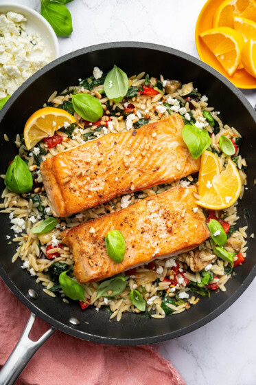 Two salmon filets in a skillet served over a bed of orzo combined with roasted red peppers, chopped dates, feta and swiss chard. It's topped with lemon slices and fresh basil.