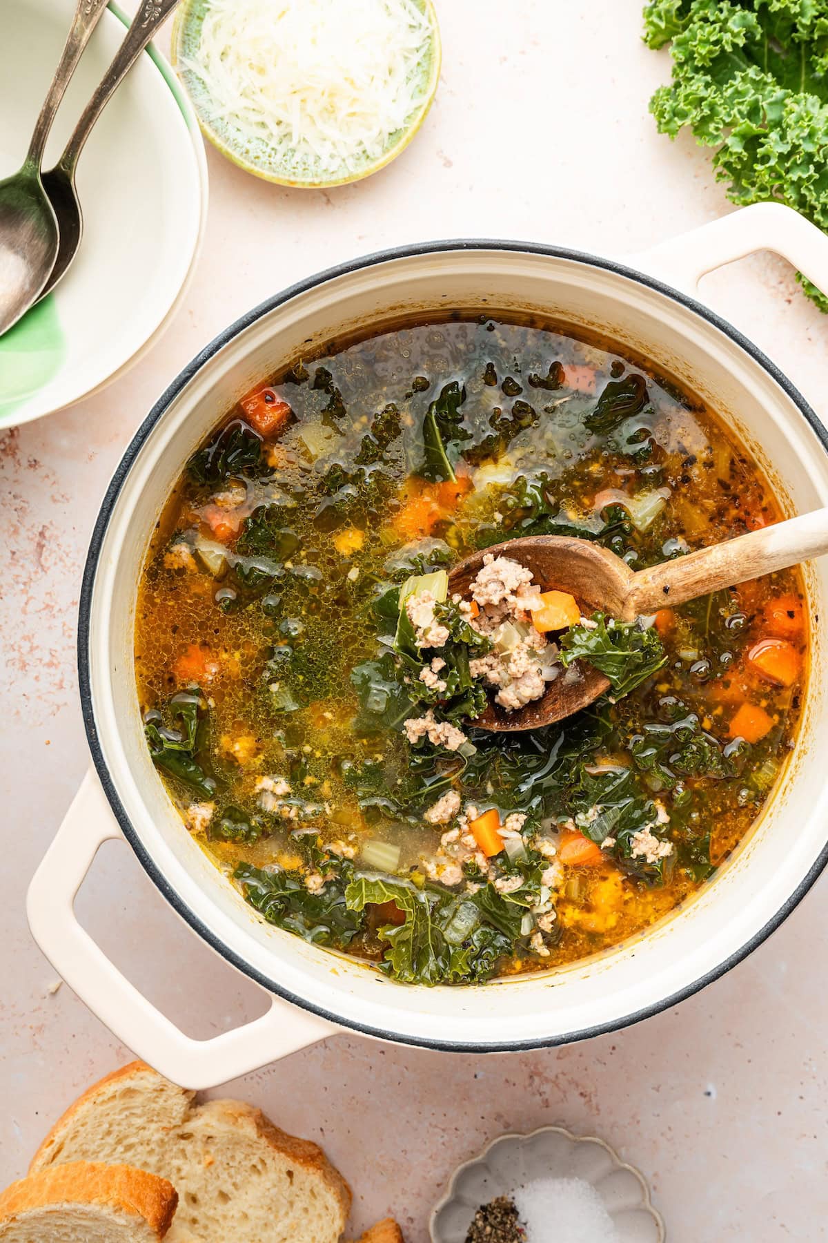 A pot of sausage and kale soup served with a wooden spoon. There is a small bowl of parmesan cheese and an empty bowl with two spoons in it next to the pot of soup.
