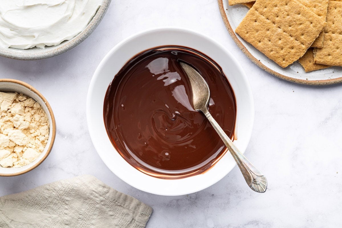 Bowl of melted chocolate with spoon, ready for layering in frozen s’mores recipe.