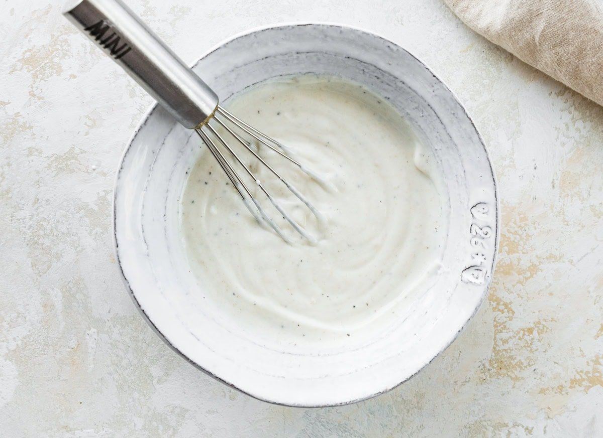 A white ceramic bowl filled with a creamy Greek yogurt sauce being whisked with a metal whisk.