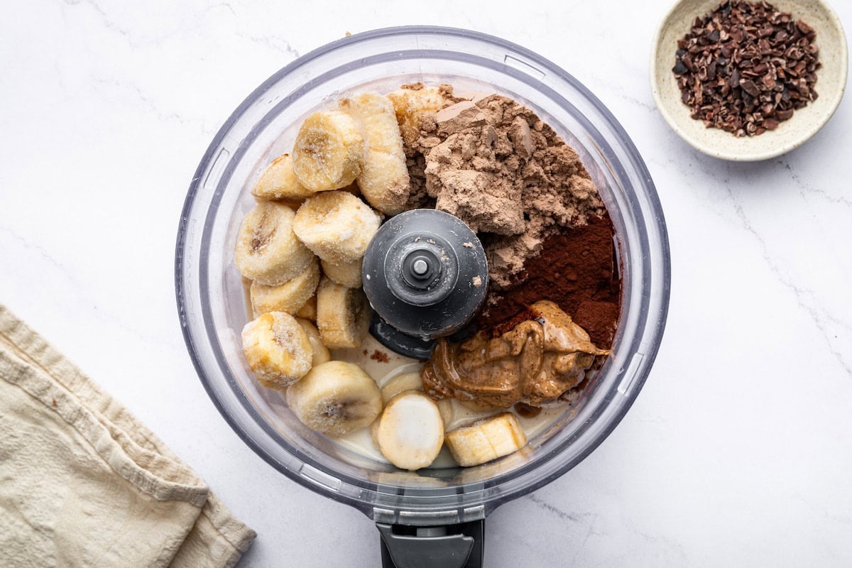 A food processor filled with frozen banana slices, cocoa powder, chocolate protein powder, almond butter, and a liquid base, ready to be blended.