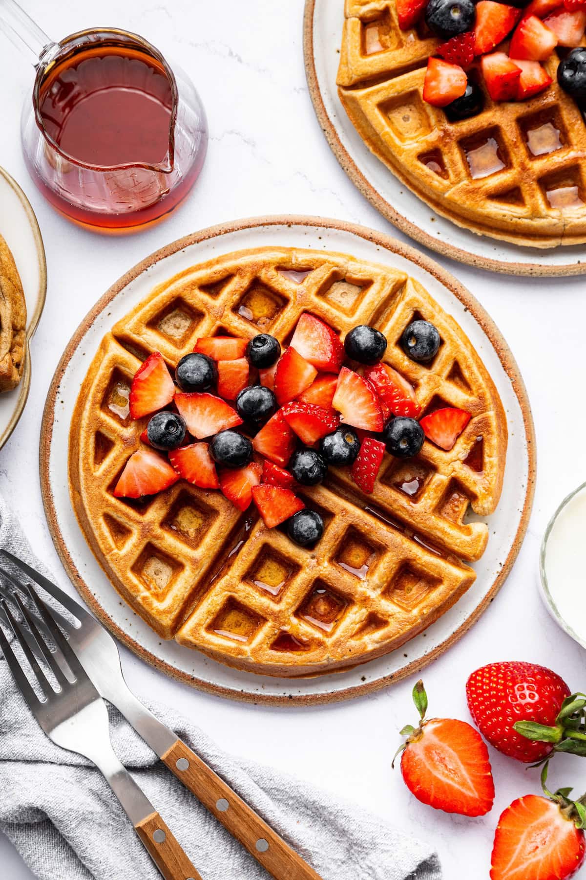 Golden brown protein waffles on a plate, topped with fresh strawberries and blueberries, drizzled with syrup, and served with a side of fruit.