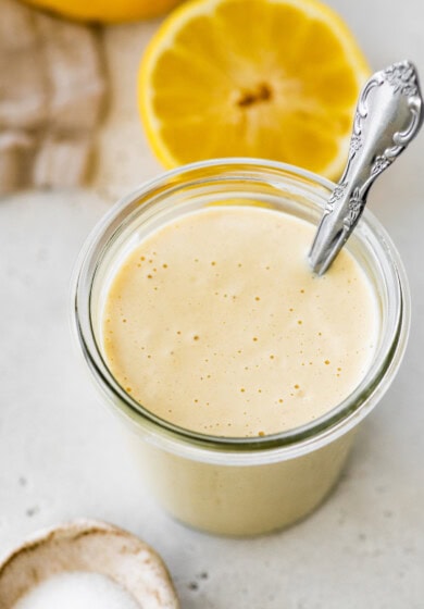 A creamy tahini dressing in a glass jar with a metal spoon and a halved lemon in the background.
