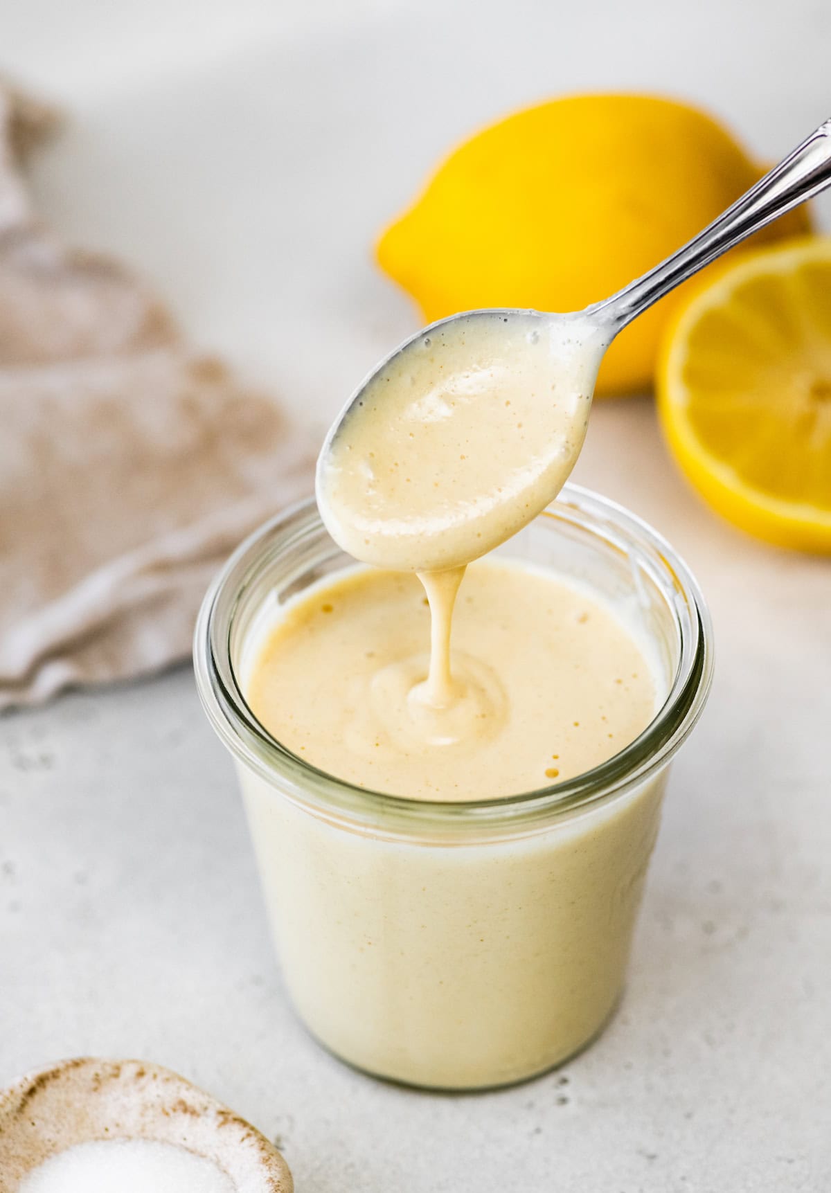 A jar of tahini dressing with a spoon lifting some of it. A lemon and a small dish of salt are in the background.
