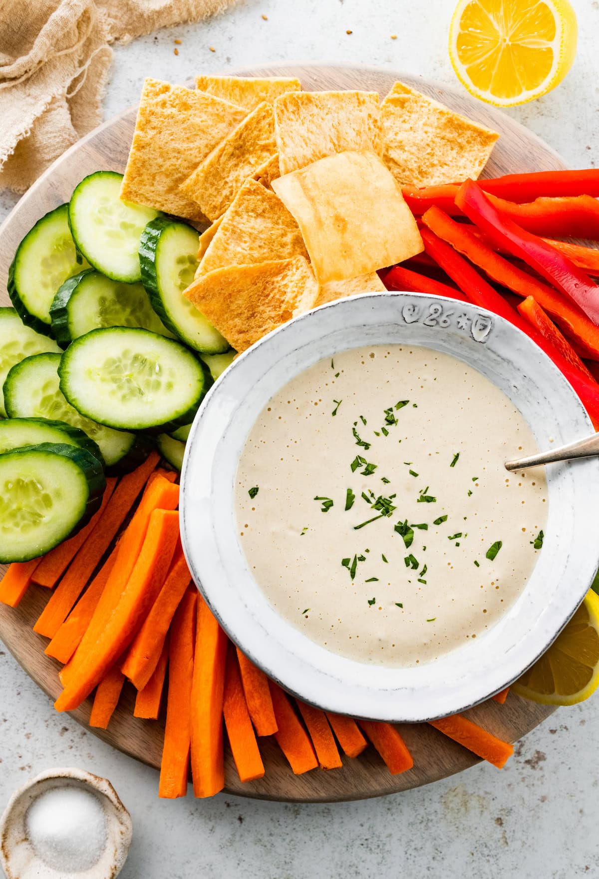 Bowl of creamy tahini sauce garnished with fresh herbs, served on a platter with sliced cucumbers, carrots, red bell peppers, and pita chips.