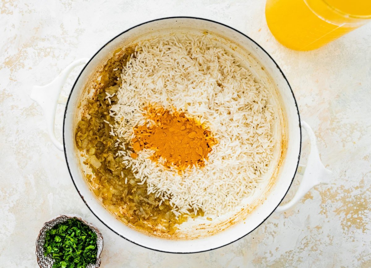 Basmati rice and ground turmeric added to a white pot with sautéed onions and garlic.