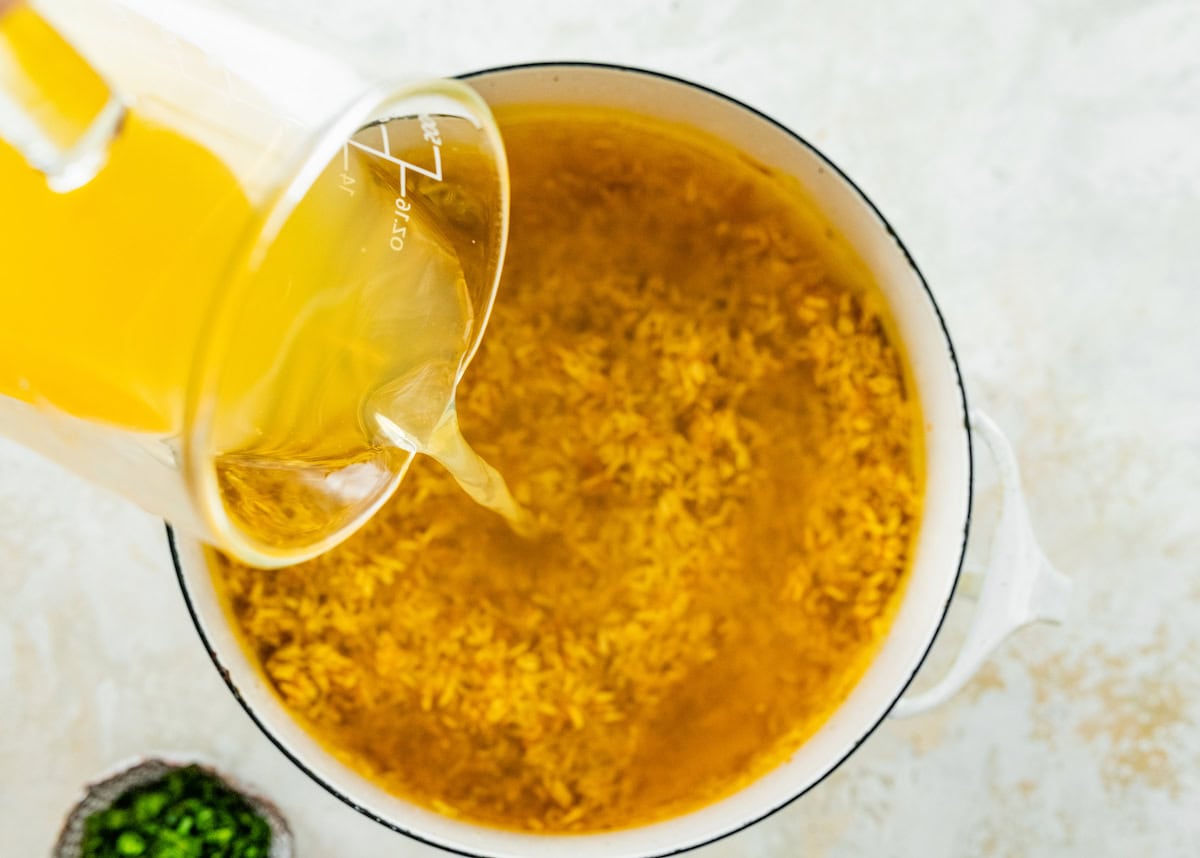 Vegetable broth being added to a white pot with basmati rice, turmeric and sauteed garlic and onions.