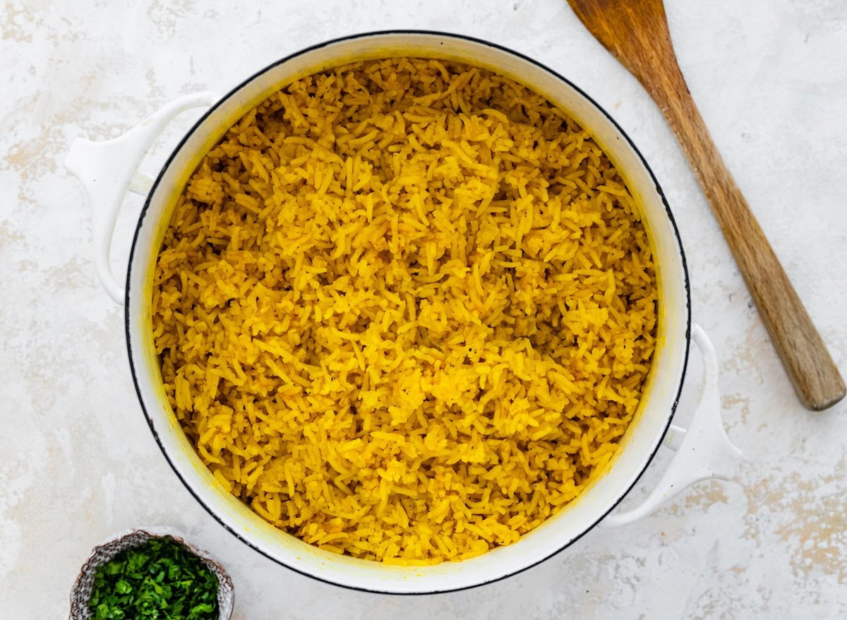 Cooked turmeric rice in a white pot. There is a wooden spoon next to the pot.