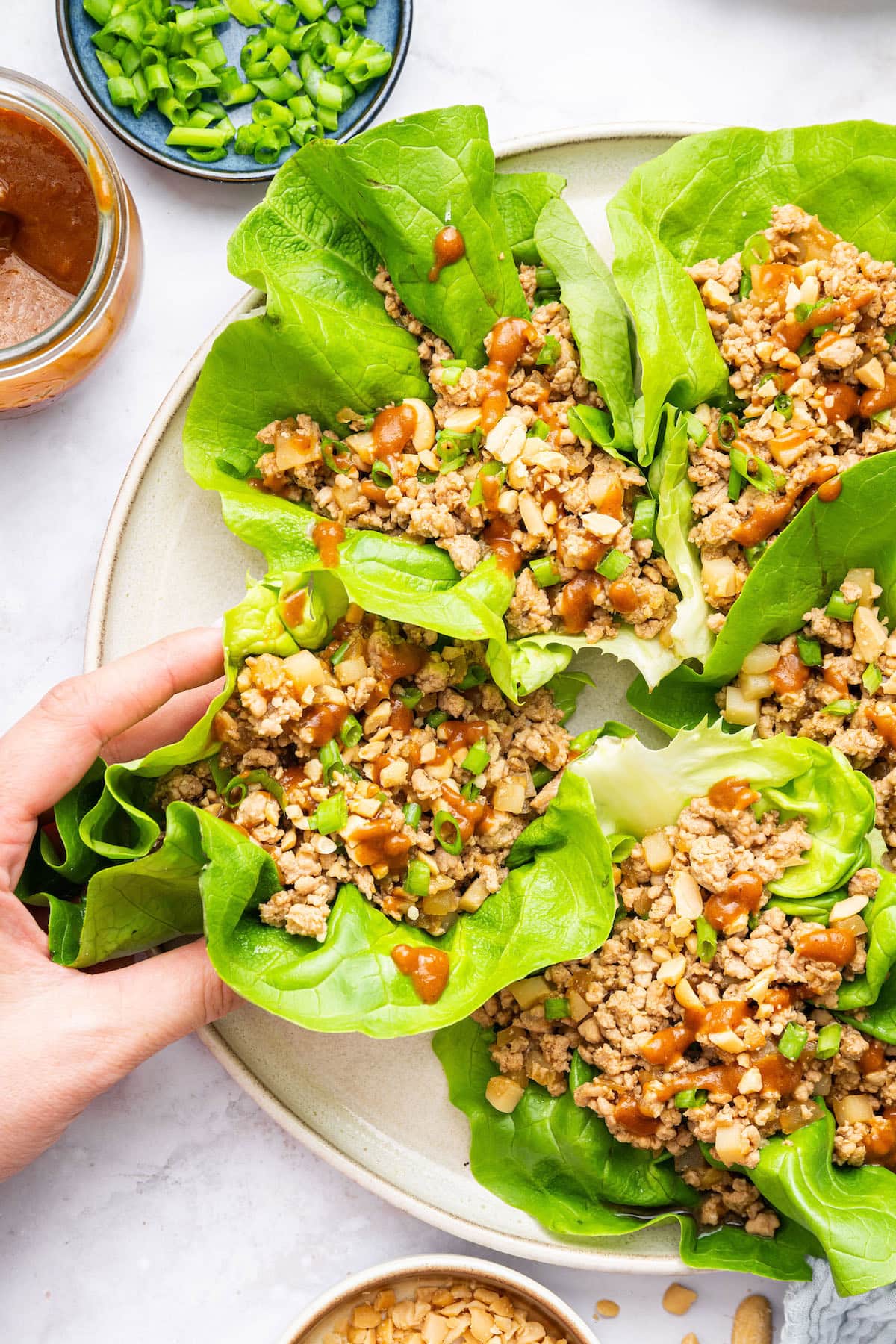 A woman's hand grabbing a chicken lettuce wrap from a plate. The lettuce wrap is topped with a peanut sauce and green onions.
