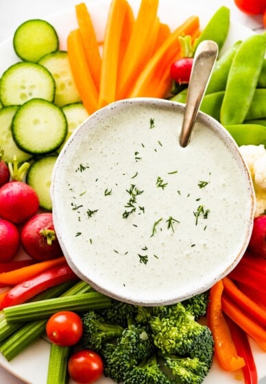 Cottage cheese ranch dip in a white bowl garnished with fresh dill. The bowl is on a plate with an assortment of fresh vegetables.