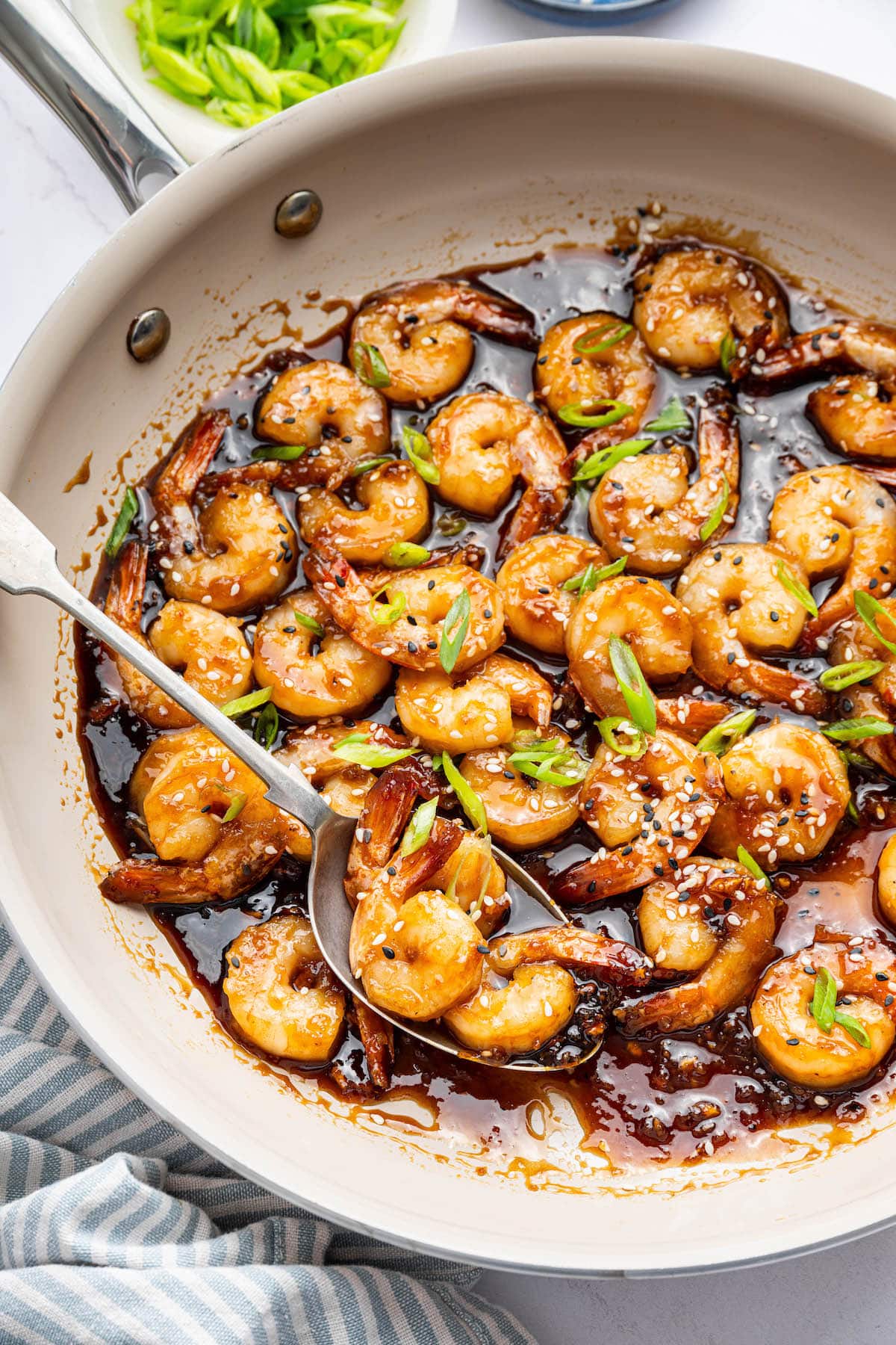 Shrimp coated in a honey garlic sauce in a skillet. There is a silver spoon in the skillet and the shrimp is topped with sesame seeds and green onion slices.