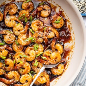 Shrimp coated in a honey garlic sauce in a skillet. There is a silver spoon in the skillet and the shrimp is topped with sesame seeds and green onion slices.