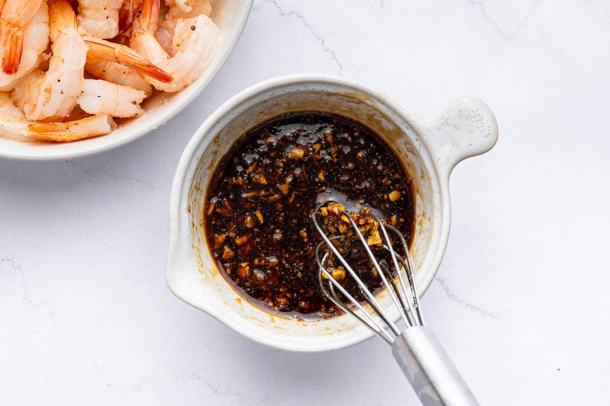 A honey garlic sauce being whisked together in a small bowl. There is a bowl with raw shrimp next to it.