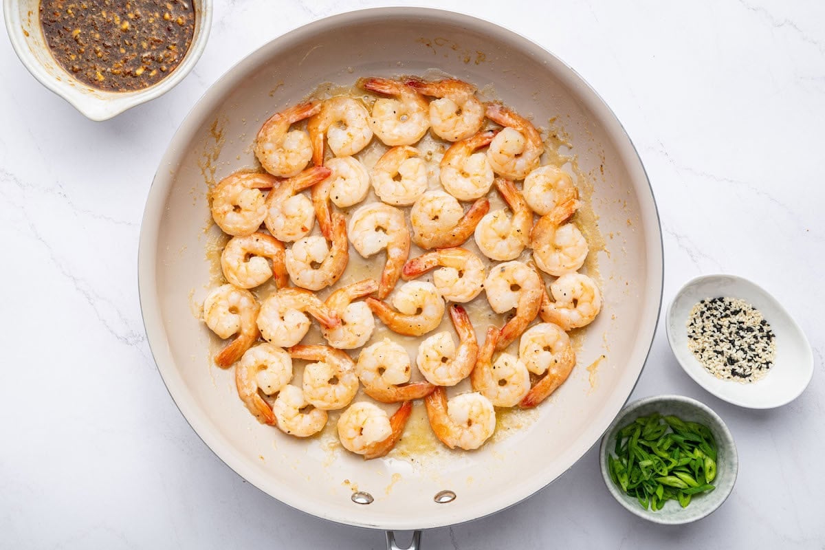 Cooked shrimp in a skillet. There are three bowls next to the skillet: one with a honey garlic sauce, one with sesame seeds and one with green onion slices.
