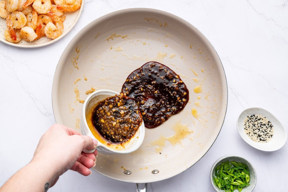A woman's hand pouring a honey garlic sauce into a skillet. There is a bowl with sesame seeds and a bowl with green onion slices next to the skillet.