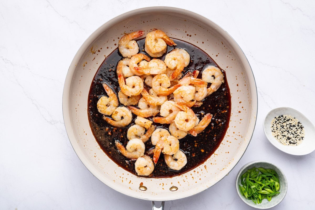 Cooked shrimp added to the honey garlic sauce in the skillet. There is a bowl with sesame seeds and a bowl with green onion slices next to the skillet.