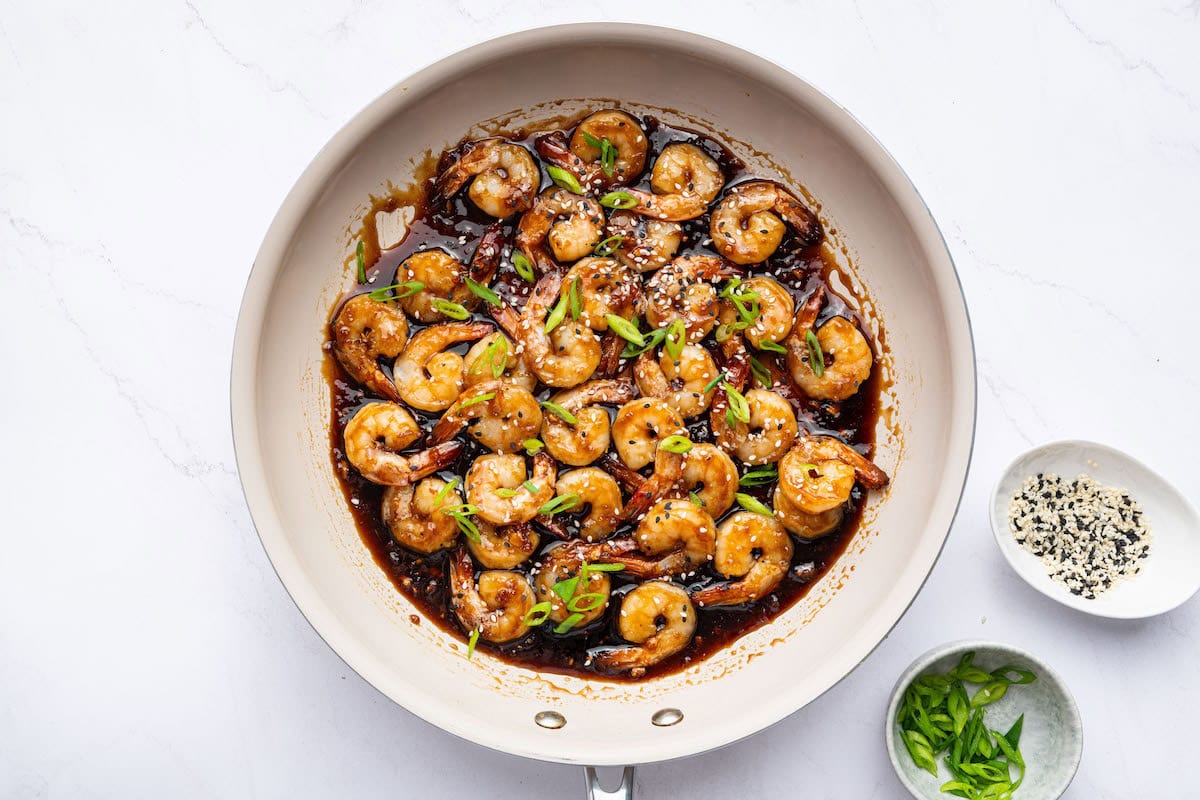 Shrimp coated in a honey garlic sauce in a skillet. The shrimp is topped with sesame seeds and green onion slices.