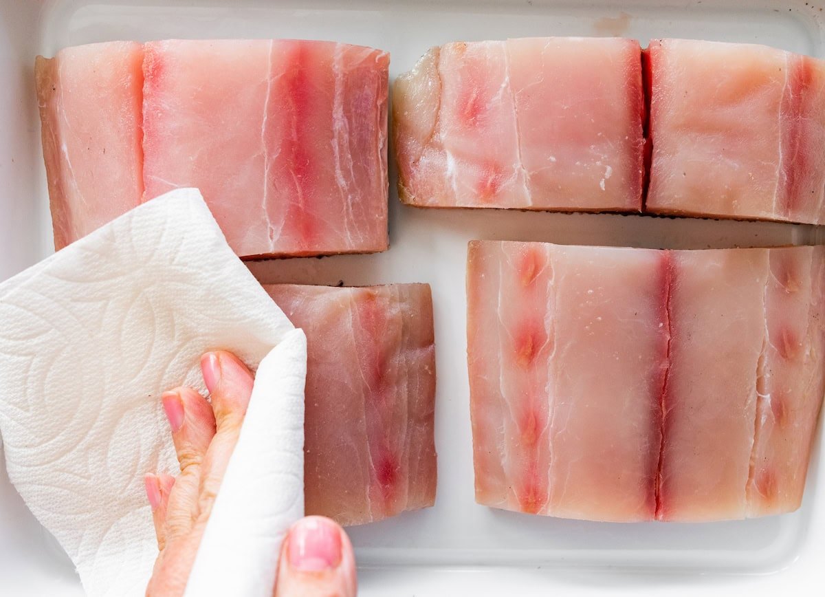 A woman's hand patting dry four mahi mahi fillets with a paper towel.