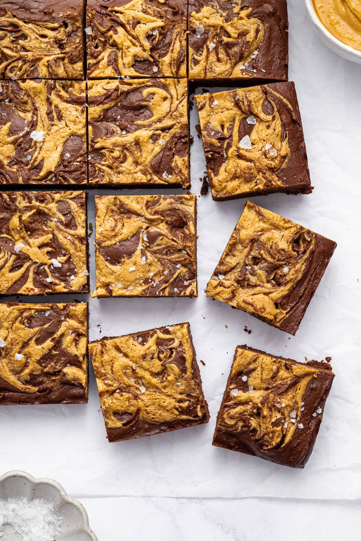 Peanut butter brownies cut into 16 squares on a marbled surface. The brownies are topped with a peanut butter swirl and flaked sea salt.