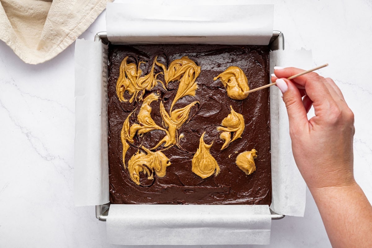 Unbaked brownie batter in a square pan with dollops of peanut butter on top. A woman's hand is swirling the peanut butter on top of the batter to get a marble look.