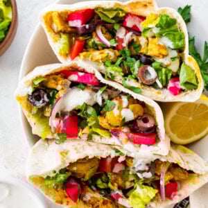Three halves of chicken shawarma pita pockets stacked in a bowl, filled with veggies, olives, pickled turnips, and topped with yogurt sauce and fresh parsley.