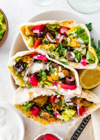 Three halves of chicken shawarma pita pockets stacked in a bowl, filled with veggies, olives, pickled turnips, and topped with yogurt sauce and fresh parsley.