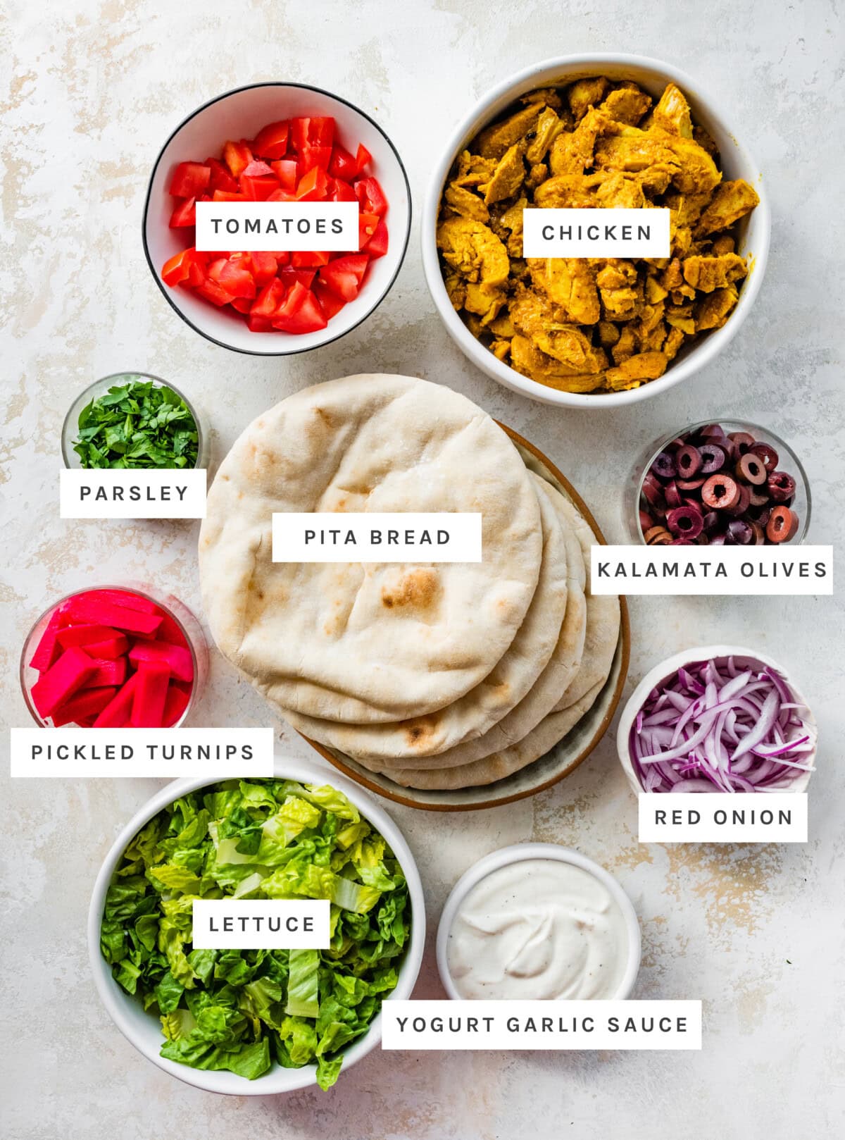 Ingredients measured out to make Pita Pocket with Chicken Shawarma: Tomatoes, chicken, parsley, pita bread, Kalamata olives, pickled turnips, red onion, lettuce, yogurt garlic sauce.