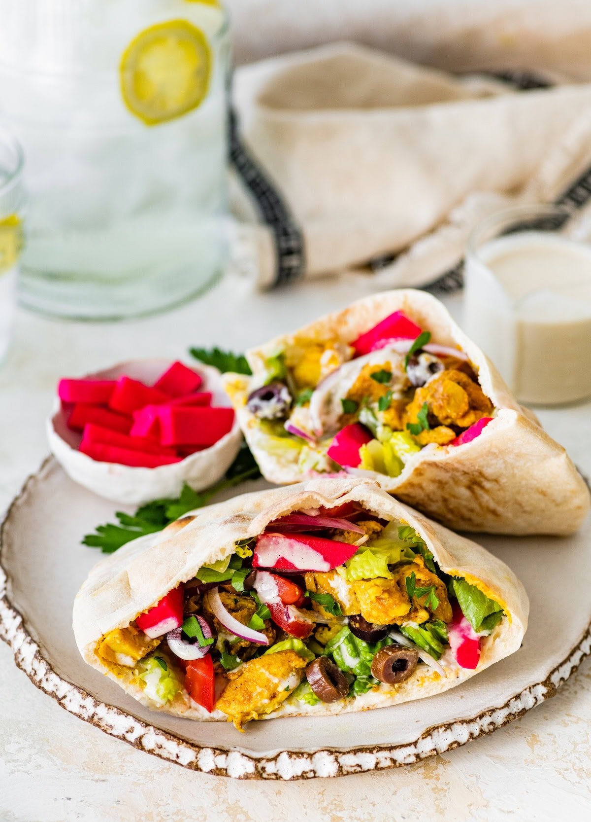 Two chicken shawarma pita pockets on a ceramic plate, surrounded by pickled turnips, Greek yogurt sauce, and garnished with fresh parsley.