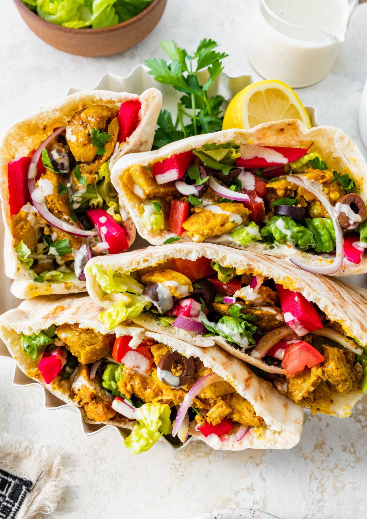 Four chicken shawarma pita pockets arranged in a white fluted serving dish, loaded with veggies, olives, pickled turnips, and yogurt sauce.