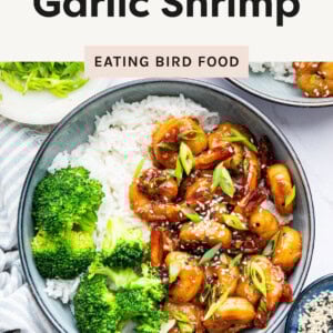 A bowl of honey garlic shrimp served over white rice, accompanied by steamed broccoli. The shrimp are coated in a glossy, caramelized sauce and garnished with sesame seeds and thinly sliced green onions. A second bowl with the same dish is partially visible in the background. A small dish of sesame seeds and a plate of additional green onions are placed nearby on a white surface with a striped cloth napkin.