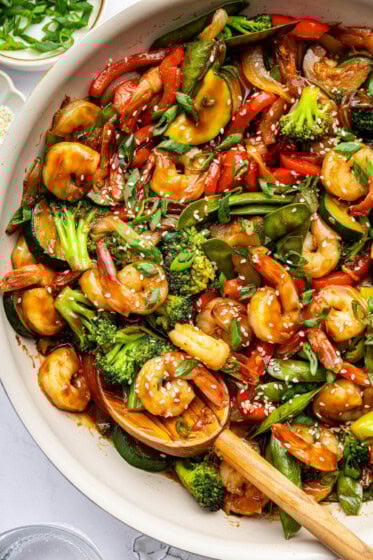 Shrimp stir-fry with shrimp, broccoli, red bell peppers, snap peas, zucchini, and onions coated in sauce. The dish is garnished with sesame seeds and chopped green onions, with a wooden spoon in the pan.