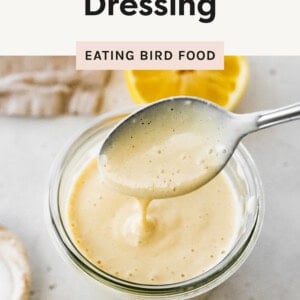 This image showcases a close-up of creamy tahini dressing in a glass jar. A spoon is lifted above the jar, allowing the smooth, thick dressing to drizzle back down, highlighting its velvety texture. In the background, there’s a whole lemon and a lemon half, suggesting the fresh, zesty ingredients used in the dressing. The neutral-toned cloth and light surface add a natural and fresh aesthetic.