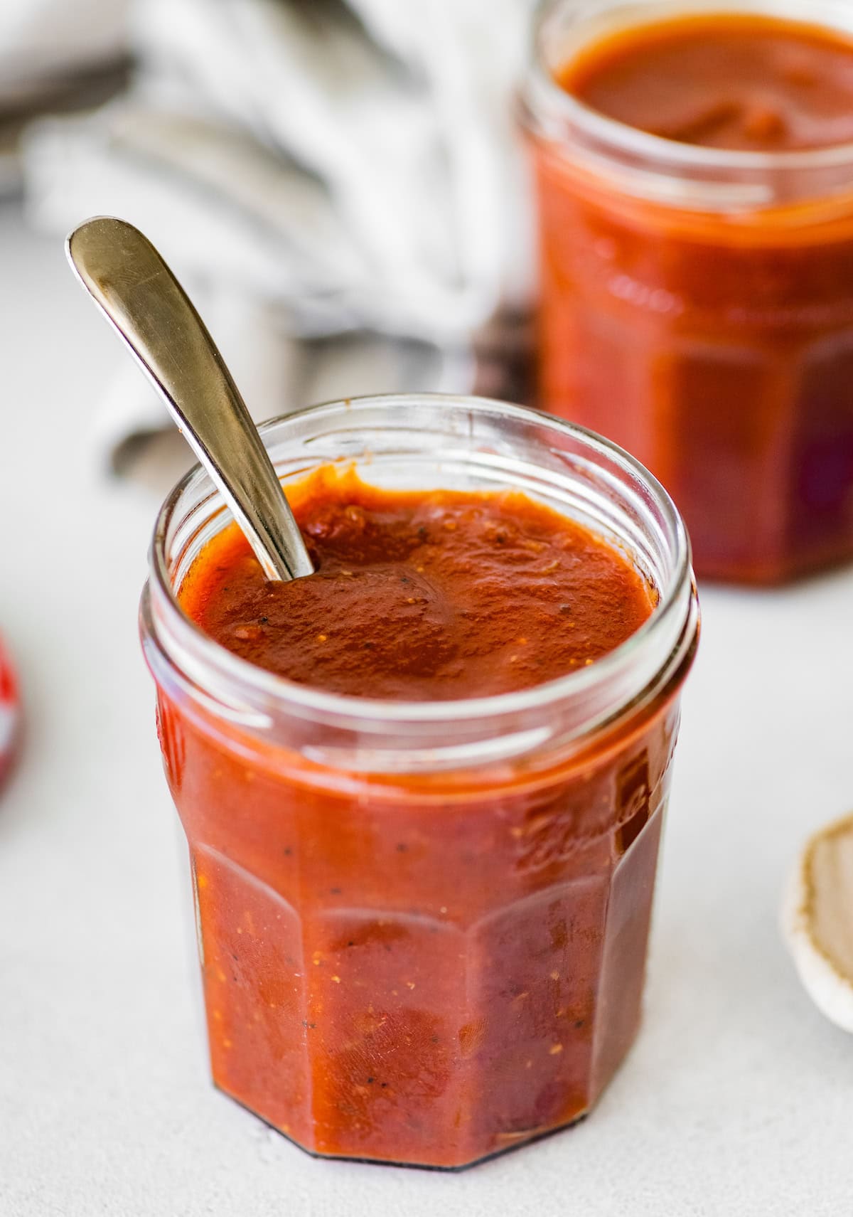 BBQ sauce in a glass mason jar with a silver spoon in the jar. There is a second glass filled with BBQ sauce in the background.