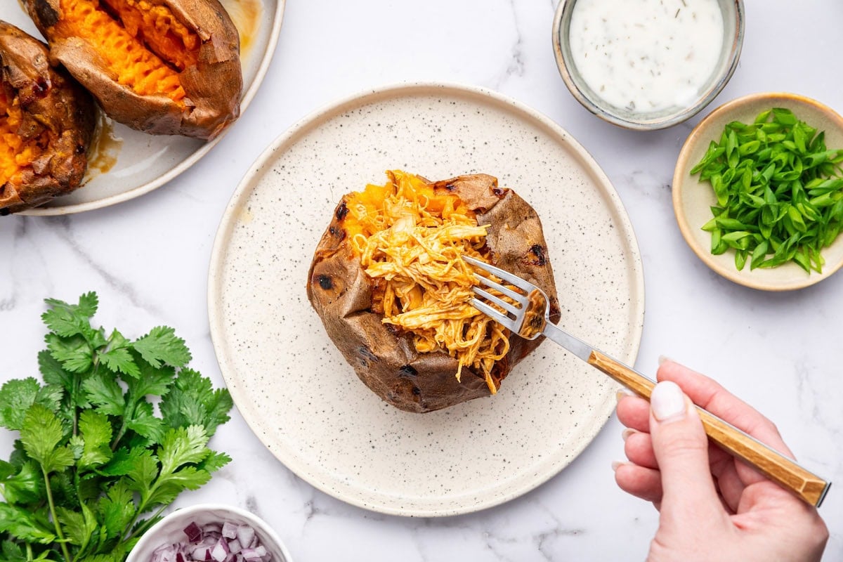 Baked sweet potato stuffed with shredded buffalo chicken and topped with ranch dressing on a white plate.