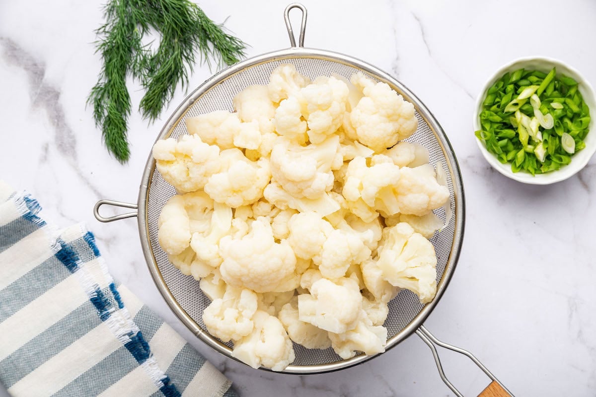 Steamed cauliflower in a colander. There is fresh dill and a small bowl with chopped green onions next to the colander.