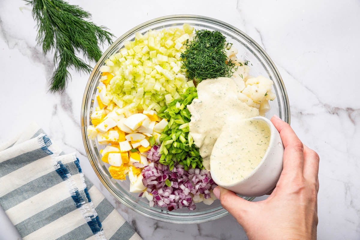 A woman's hand pouring the creamy mayo dressing over the rest of the cauliflower potato salad ingredients in a large glass bowl.