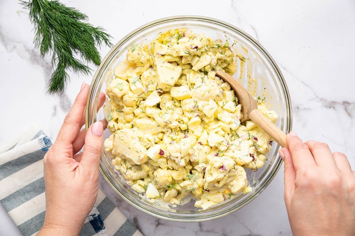 A woman's hand mixing together the cauliflower potato salad in a marge glass mixing bowl with a wooden spoon.