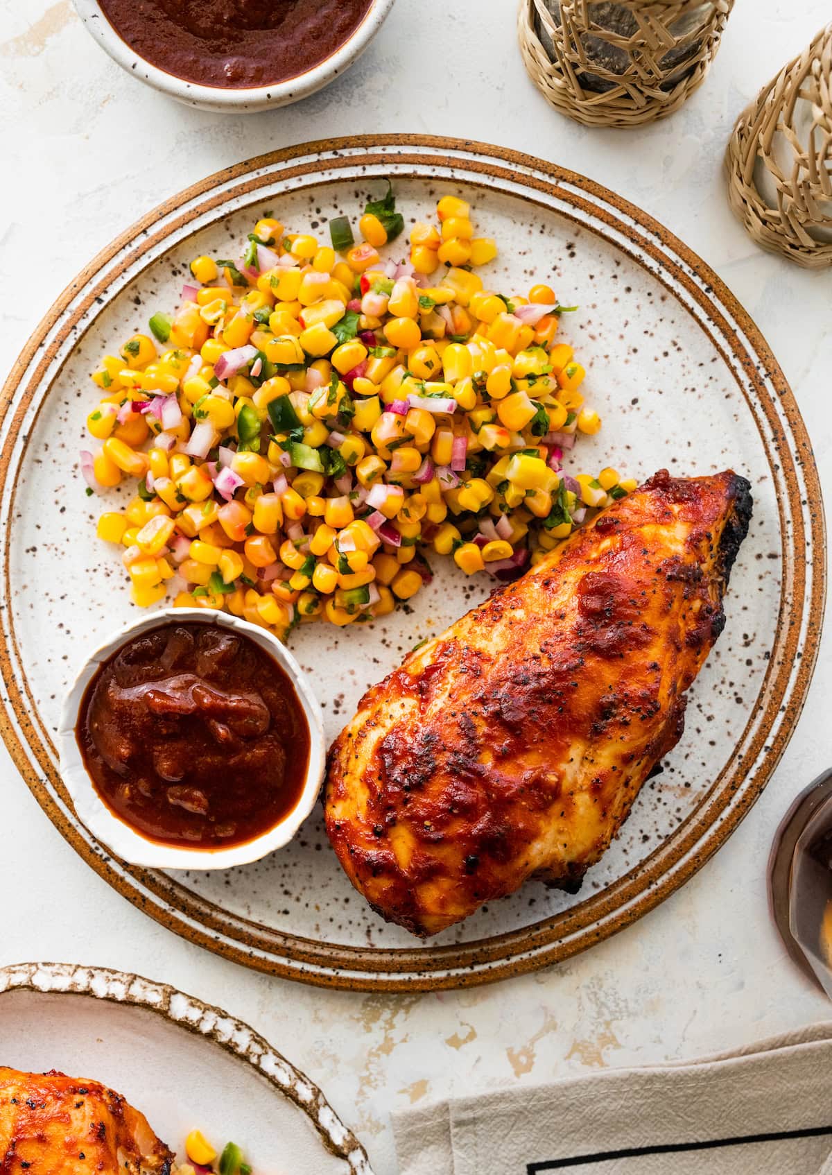 One grilled BBQ chicken breast on a plate served with a side of BBQ sauce and a corn salad.