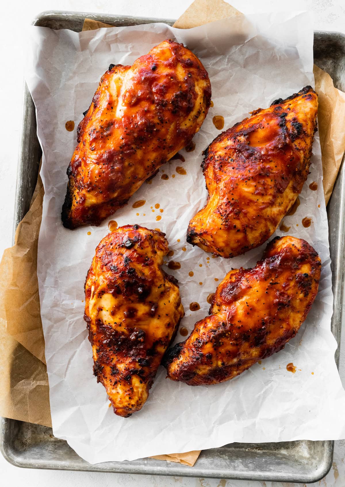 Four grilled BBQ chicken breasts on a small baking sheet lined with parchment paper.