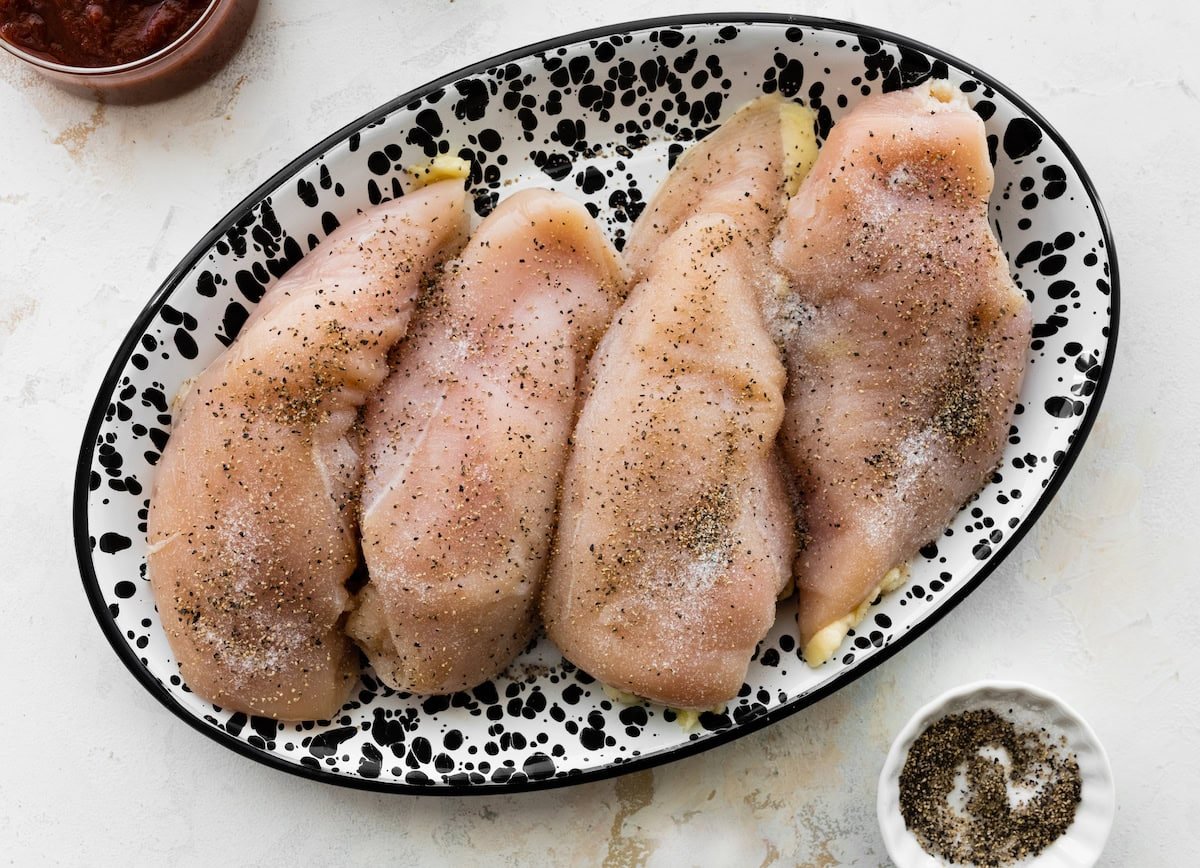 Four raw chicken breasts on a serving plate seasoned with salt and black pepper.