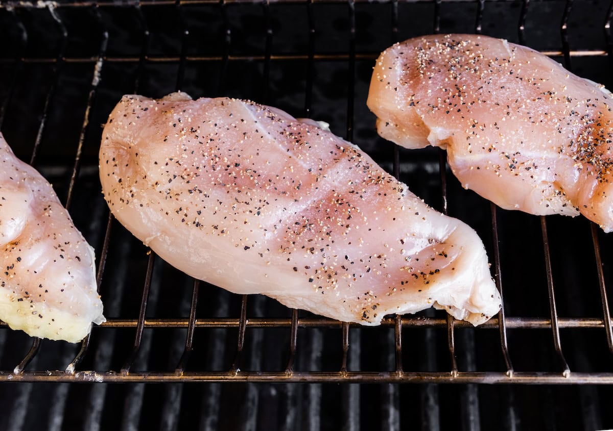 Three raw chicken breasts cooking on a grill grate. The chicken breasts are seasoned with salt and pepper.
