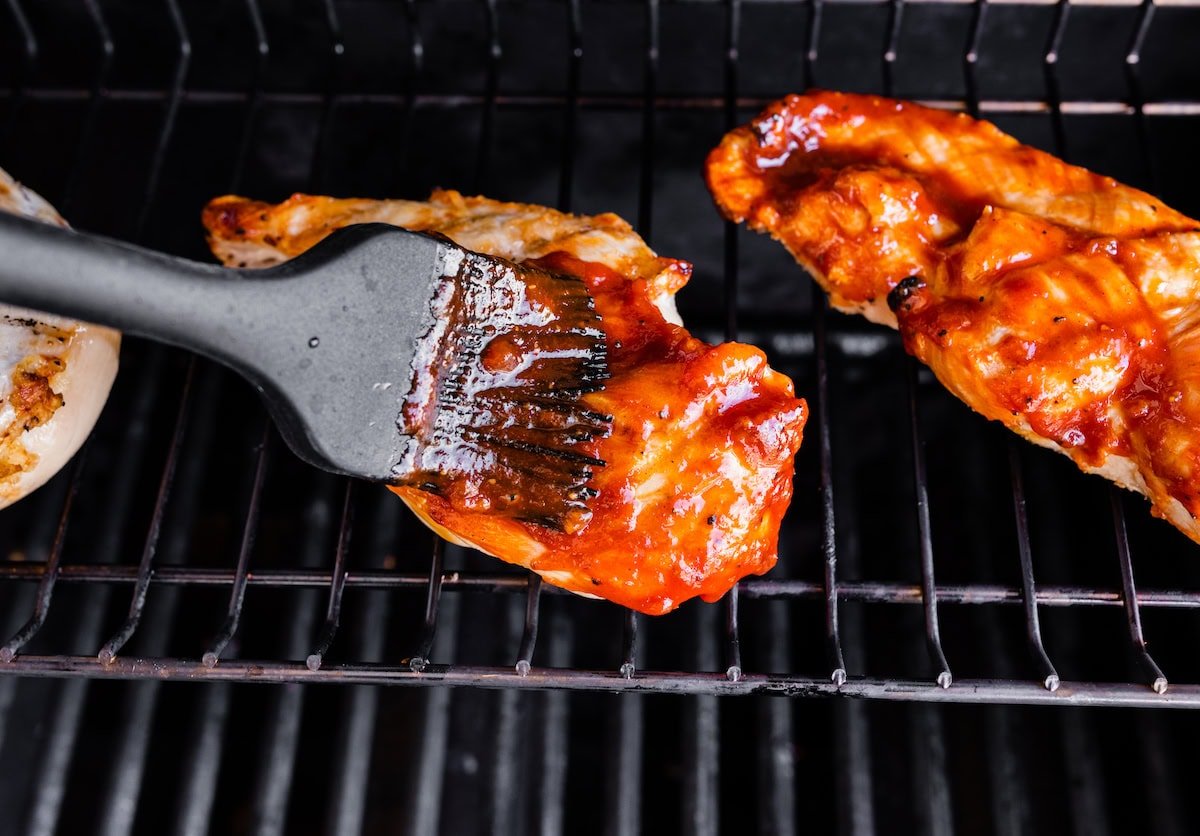 A silicone basting brush brushing BBQ sauce on the cooked chicken breasts on the grill grates.