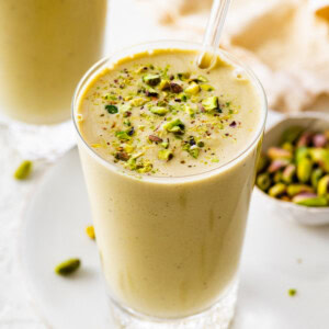 One glass filled with a pistachio smoothie topped with crushed pistachios. There is a clear straw in the glass and another smoothie in the background.