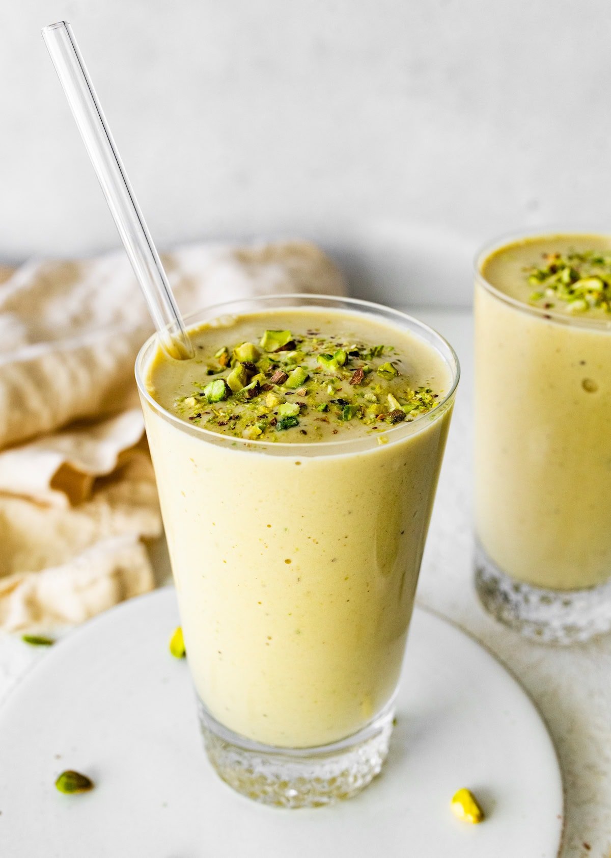 Two pistachio smoothies in glasses topped with chopped pistachios. The front glass has a clear straw in it.
