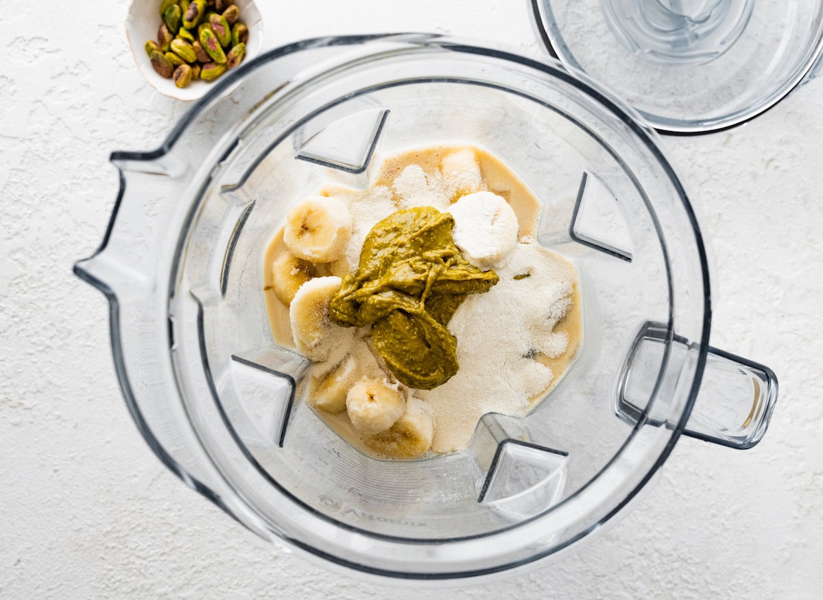 Ingredients for the pistachio smoothie in the base of a blender: almond milk, frozen banana, pistachio butter, vanilla protein powder and ice.