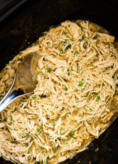 Shredded salsa verde chicken in a slow cooker with a silver serving spoon. Fresh cilantro is on top of the chicken.