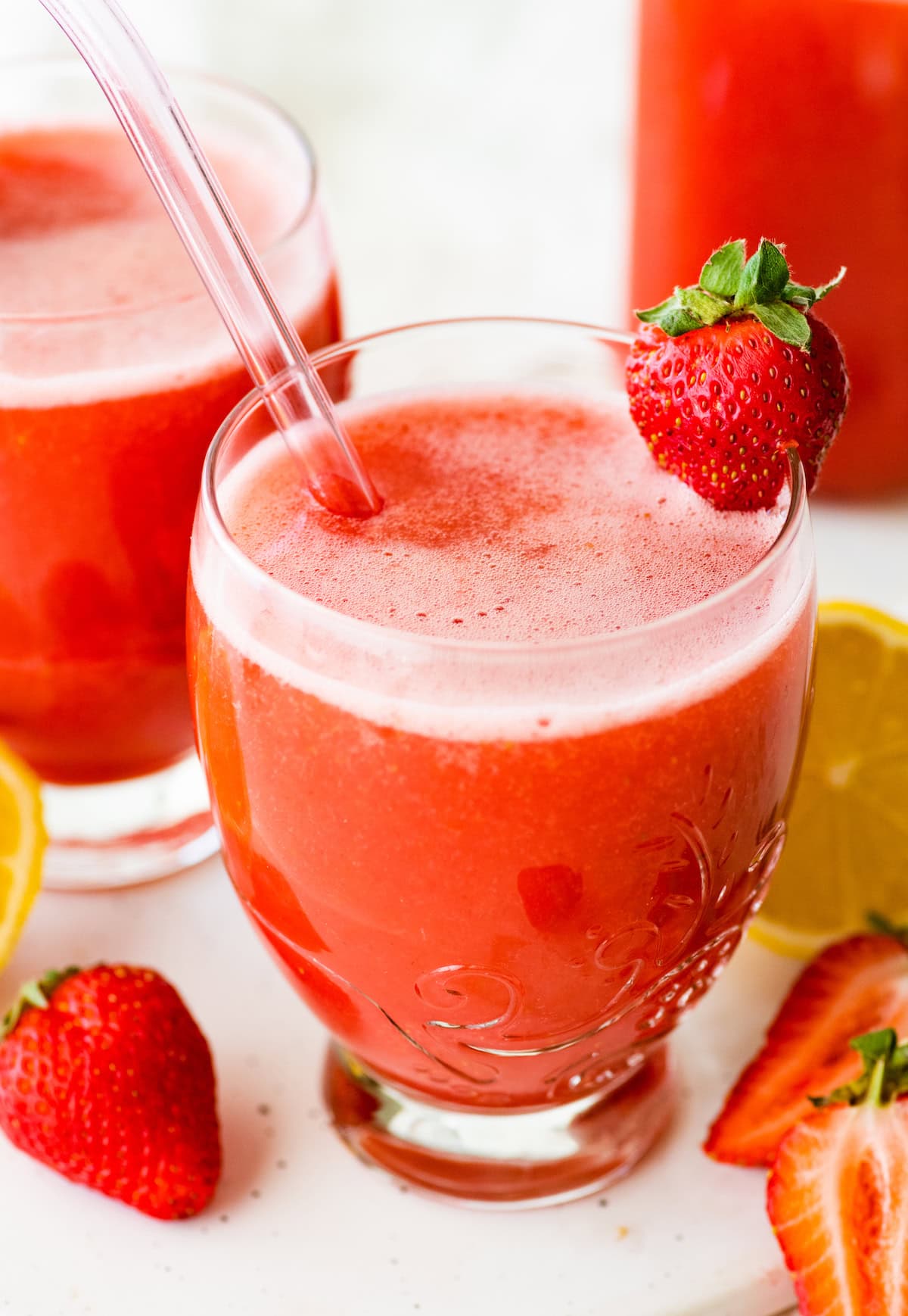 Glass of strawberry juice with a glass straw and a fresh strawberry garnish, with another filled glass and fruit in the background.