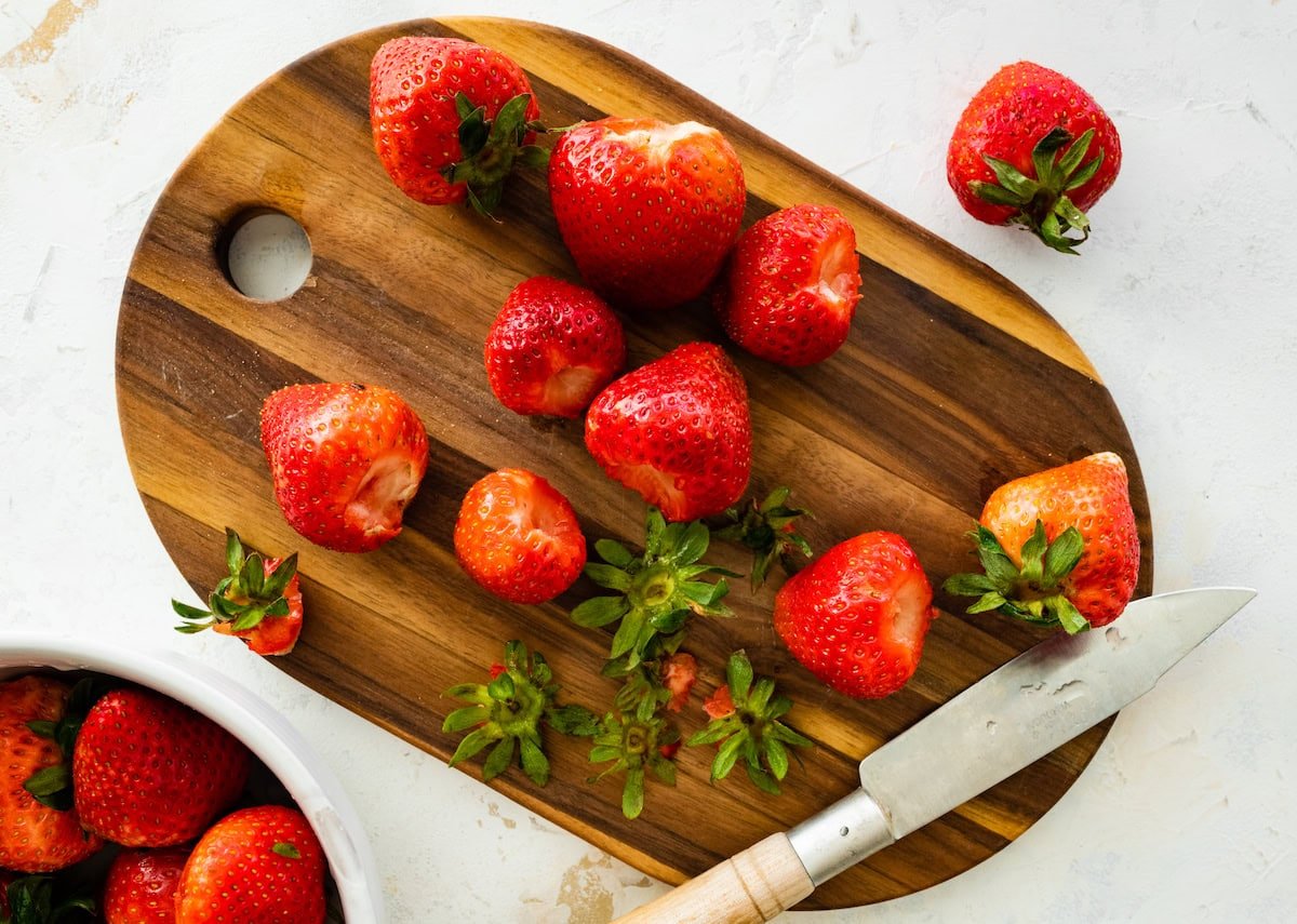 Chopped strawberries on a wooden cutting board with green tops removed, a paring knife, and a bowl of whole strawberries nearby.