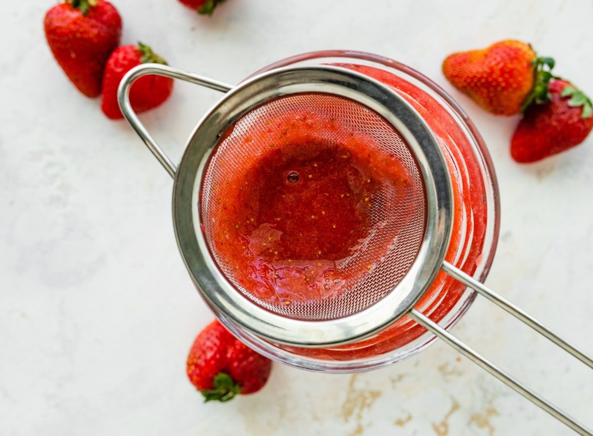 Strawberry puree being strained through a fine mesh sieve over a glass jar, with pulp collecting in the strainer.