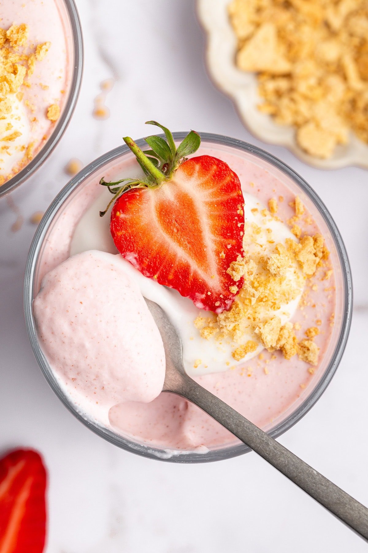 One glass filled with high-protein strawberry mousse topped with a dollop of cocowhip, one halved fresh strawberry and crushed graham crackers. There is a spoon scooping out a bite from the glass.